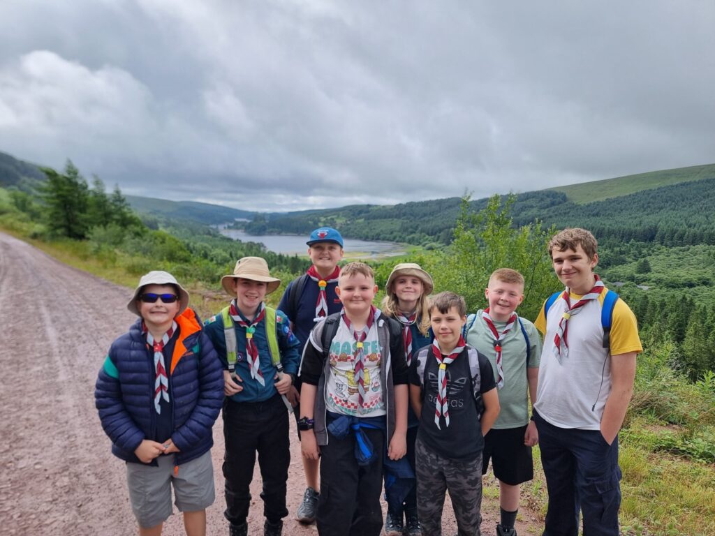 A group of Scouts mid-hike posing for a photo. the background shows a lake in the distance between hills to the left and right.